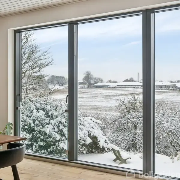 Stort glasparti med sneklædt landskab udenfor. Træer og marker dækket af sne ses gennem vinduerne i en lys stue med spisebord og stole.