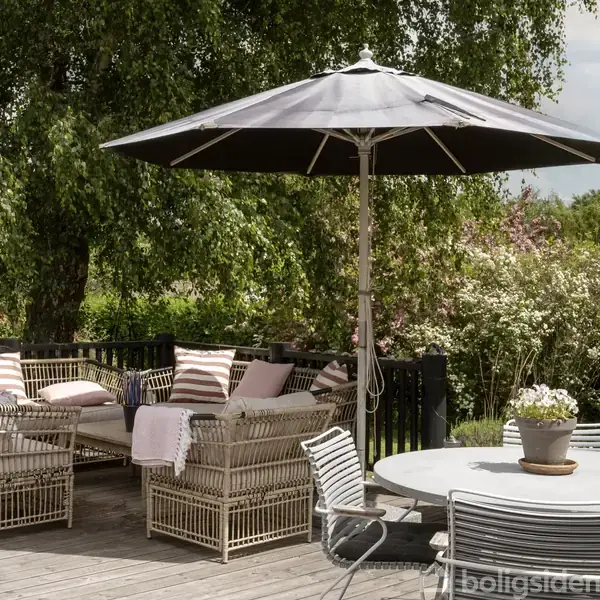 Terrassemøbler med flettede sofaer og puder står under en stor parasol. Omgivet af grønne træer og buske, skaber det en hyggelig udendørs opholdsstue i en have.