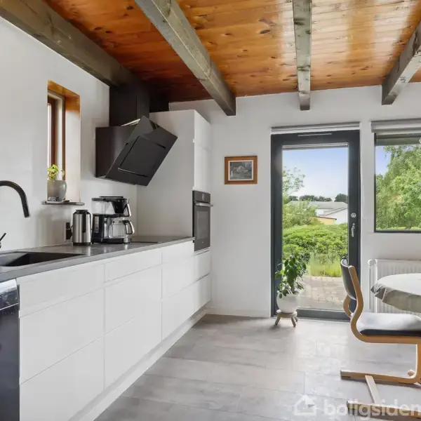 Køkken med hvidt skabsarrangement, sort vask og kogeplade. Træloft og fritlagte bjælker. Rundt spisebord med stole ved vindue, udsigt til have gennem glasdør og vindue.