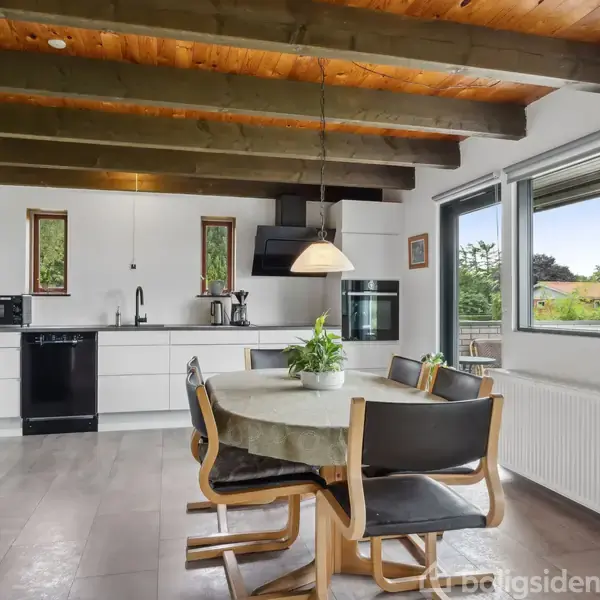 Et spisebord med fire stole står under en lampe. Bagved ses moderne køkkenelementer med hvide skabe og sorte apparater. Stort vindue med udsigt til grøn have.