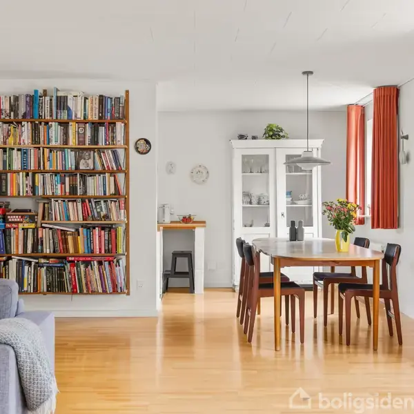 Bogreol fyldt med bøger ved siden af en grå sofa i en stue. Spisebord med stole og vase med blomster står ved vinduer med røde gardiner. Klokke på væggen.