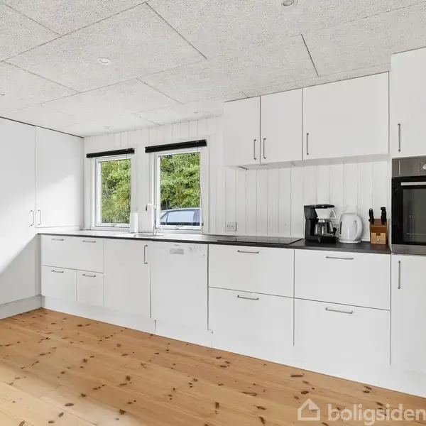 Et moderne køkken med hvide skabe og sort bordplade. Høj ovn indbygget. Dagslys strømmer ind gennem to vinduer, der viser grønne omgivelser udenfor. Trægulv og minimalistisk indretning.