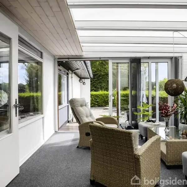 Værelset har møbler af flet, der står omkring et sofabord med planter og dekoration. Store vinduer og døre slipper lys ind og afslører en terrasse og grønne omgivelser udenfor.