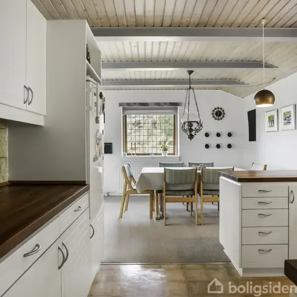 Køkkenet har hvide skabe og træbordplader, og der er en spiseplads med træstole og en lysekrone i baggrunden. Vægdekorationer og vindue fuldender rummet.