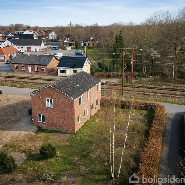Rødt murstenshus står ubenyttet i en grønt område nær jernbanespor. Omgivet af beboelsesejendomme og træer, ses en vej i nærheden, der fører til et boligområde.