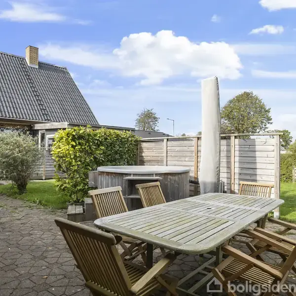 En have med et stort træbord omgivet af stole står under åben himmel. I baggrunden ses et halvtag, grønne hække og et træhus med et mørkt tag.