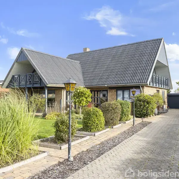 Et moderne, to-etagers hus med skrå tag beklædt med grå tagplader, flankeret af grønne buske og en brostensbelagt indkørsel med lamper, i et roligt, beboelsesområde under en blå himmel.