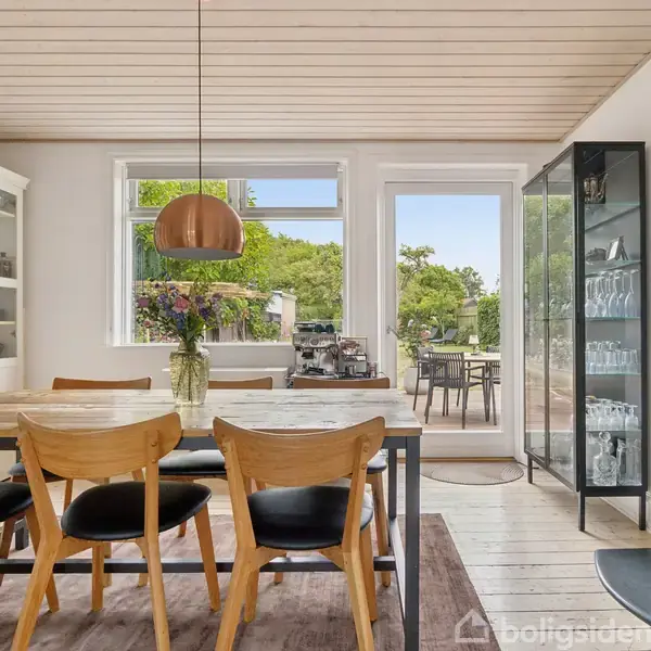 Spisebord med træstole står i lys spisestue. Vase med blomster på bordet. Glasvitrine ved siden af brændeovn. Dør og vinduer åbner ud mod terrasse og have.