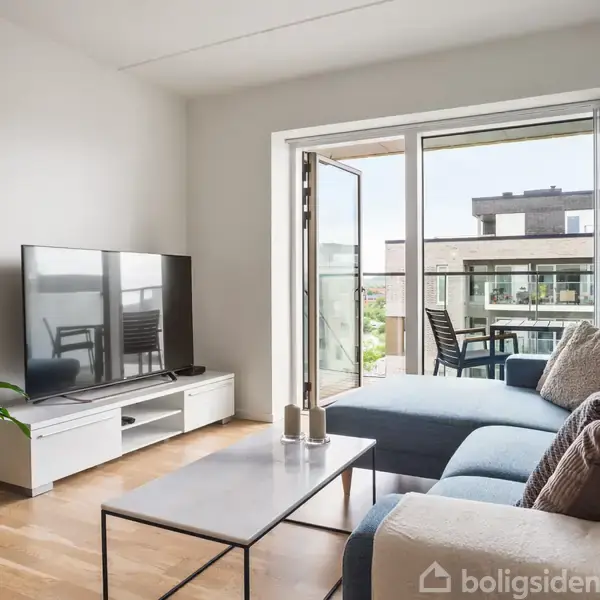 En stue med en blå hjørnesofa og sofapuder, står foran en fladskærms-TV på et lavt skab. En altandør åbner ud til en terrasse med udsigt over nabobygninger. En grøn plante er placeret ved siden af TV'et.