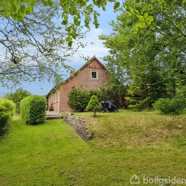 En rød murstensbygning står i en frodig have omkranset af høje træer. En græsplæne fører op til huset, mens en træbelagt terrasse er synlig i baggrunden.