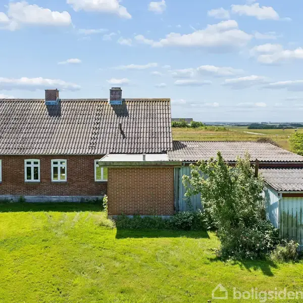 Et rødt murstenshus med gråt tegltag står stille på en grøn græsplæne. Ved siden af er en mindre tilbygning. Omgivet af træer og åbent landskab under en blå himmel.