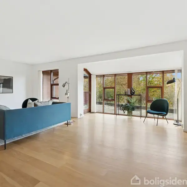 En stue med en blå sofa står på et trægulv, omgivet af lysindfald fra store vinduer. Rummet indeholder også en sort lænestol, en gulvlampe og vægkunst.