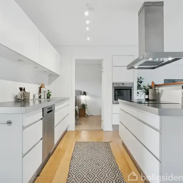 Hvidt moderne køkken med to parallelle bordplader. Midtgangen har et dekorativt tæppe. I baggrunden er der en åbning til et andet rum med stearinlys og planter på bordet.