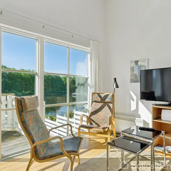 To lænestole står ved siden af et stort vindue med udsigt til en terrasse og skov. Et tv står på et møbel i hjørnet. Sollys strømmer ind.