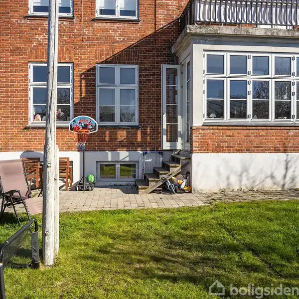 En murstensbygning med store vinduer står i solen. En lille terrasse har møbler, legetøj og en basketballkurv. Græsplænen omgiver området med en skyfri himmel i baggrunden.