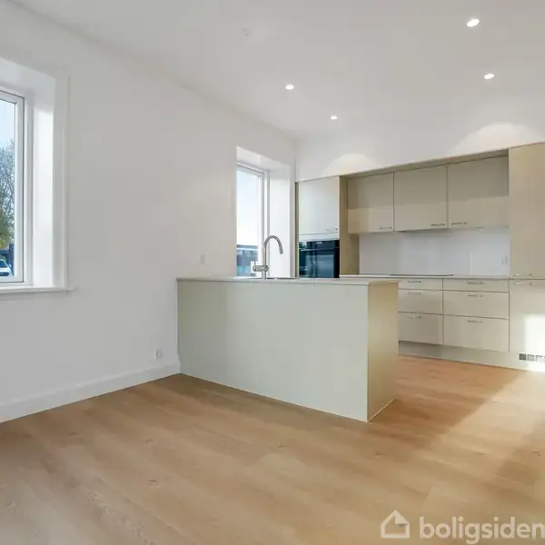 En moderne køkkenø står centralt uden aktivitet i et lyst rum med trægulv, indbyggede skabe og vinduer, der giver udsigt til en grøn have.