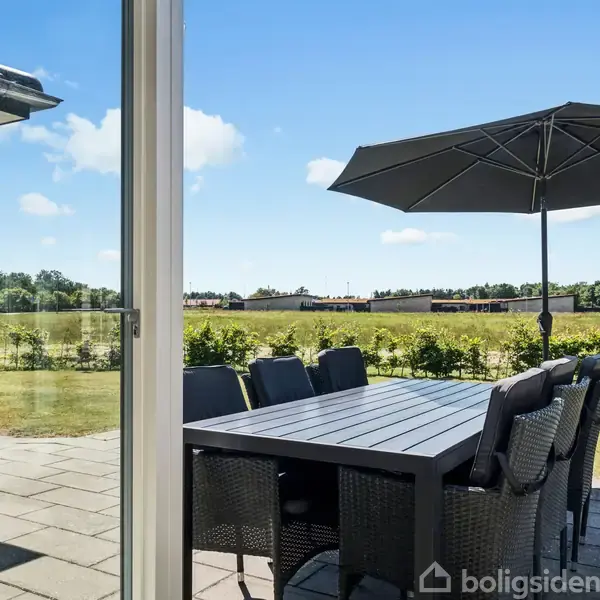 Terrassemøbler står på en flisebelagt terrasse med en sort parasol opslået over et bord og stole. I baggrunden ses en grøn mark og klar blå himmel.