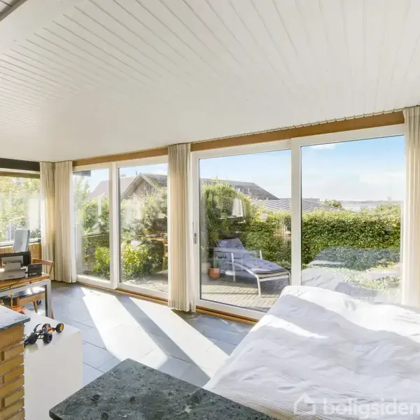 Soveværelse med seng foran store vinduer. Sollys strømmer ind, afslørende en terrasse med stol og ubeskrivelig udsigt. Planter og møbler pryder rummet, skaber en lys og åben atmosfære.