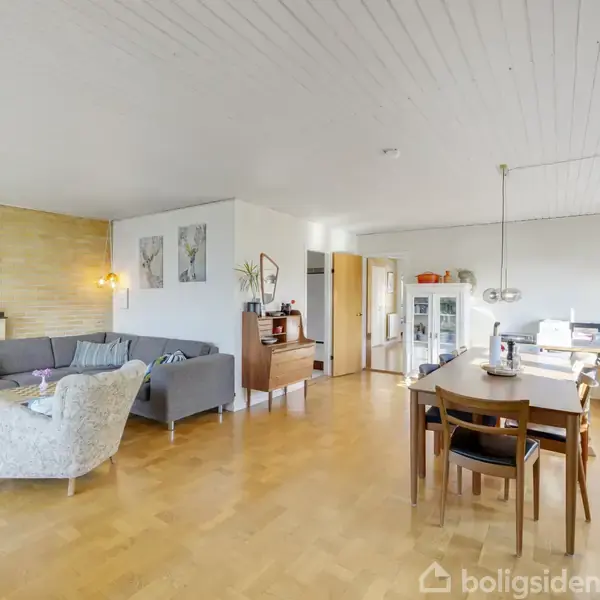 Stue med sofa- og spisearrangement. Sofa i gråt ved stenvæg med malerier, bord, lænestol. Spisebord med stole, lampe over, nær vindue. Trægulv, lyse vægge, naturligt lysindfald.