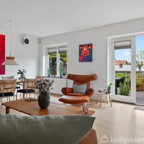 En lys stue med et stort vindue og åben dør til terrasse. Brunt lænestol og sofabord i forgrunden, spisebord med stole og rød kunst hængende på væggen.