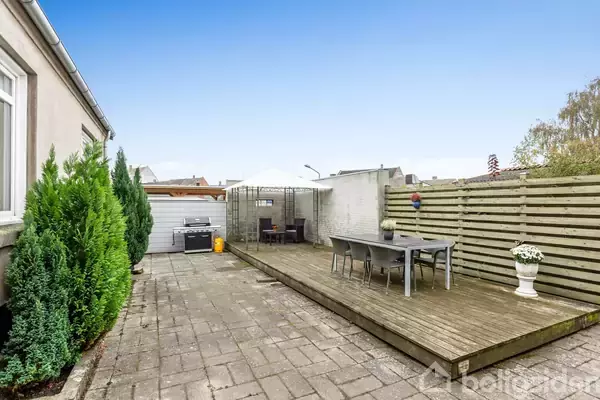 Terrasseområde med et bord og stole på et hævet trægulv mod en trælåge. Til venstre ses en grill ved siden af grønne buske.