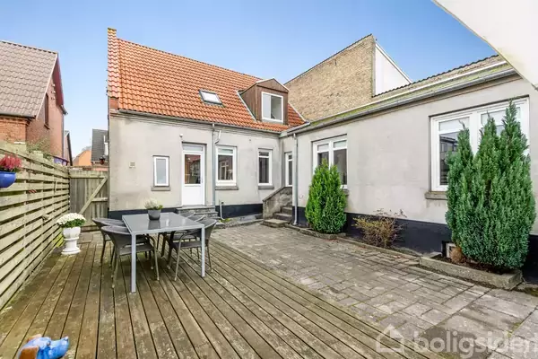 Et bord med stole står på en træterrasse. Huset har beige vægge og rødt tegltag. To buske pryder fliseområdet, omgivet af hegn og nabohuse.