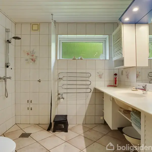 Badeværelse med bruser og toilet, flisevægge med blomsterdetaljer, langt hvidt bord med spejl og skuffer. Vinduet giver naturligt lys, og en skammel står på gulvet.