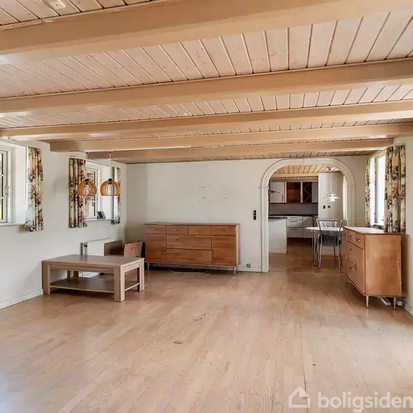 Stue med trægulv og træloft. To skænke og sofabord står opstillet. Store vinduer med blomstrede gardiner slipper lys ind. En bueåbning fører til et tilstødende rum. Ingen tekst.