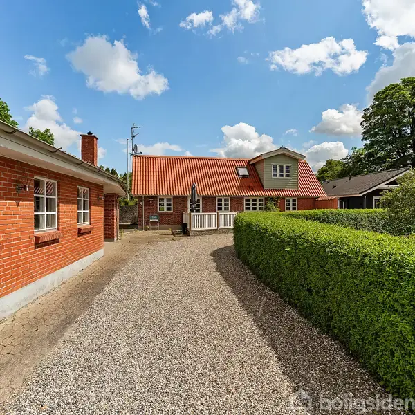Et rødt murstenshus med rødt tegltag står langs en grusvej, hvor en grøn hæk afgrænser vejen. Baggrunden er en klar blå himmel med spredte skyer.