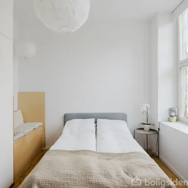 Soveværelse med en dobbeltseng centreret mod en lys væg. Til højre står et lille bord ved et vindue, hvor lys skinner igennem. Enkel, moderne indretning.