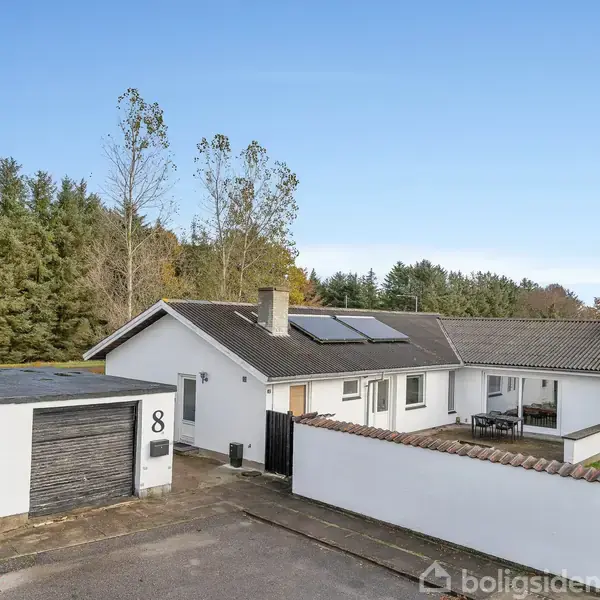 Hvidt hus med gråt tag og solpaneler står i forgrunden. Foran huset ligger en garage med tallet “8”. Omkring er der grønt græs og høje træer i baggrunden.