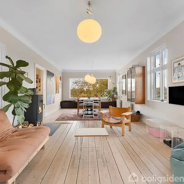 En moderne stue med lys indretning, træmøbler og en behagelig sofa. Store vinduer giver rigeligt lys, og der er kunst på væggene. En stor plante står ved siden af sofaen.