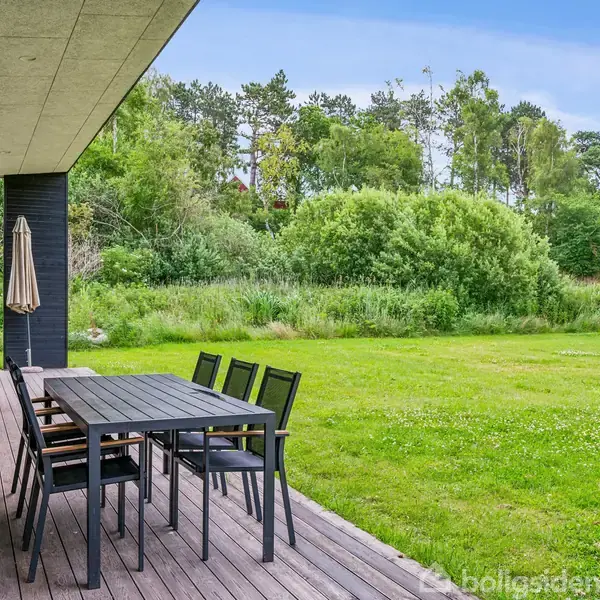 En sort spisebordssæt med otte stole står på en træterrasse ved siden af et hus. En parasol er ved enden af bordet. Omkring er der en græsplæne med træer i baggrunden.