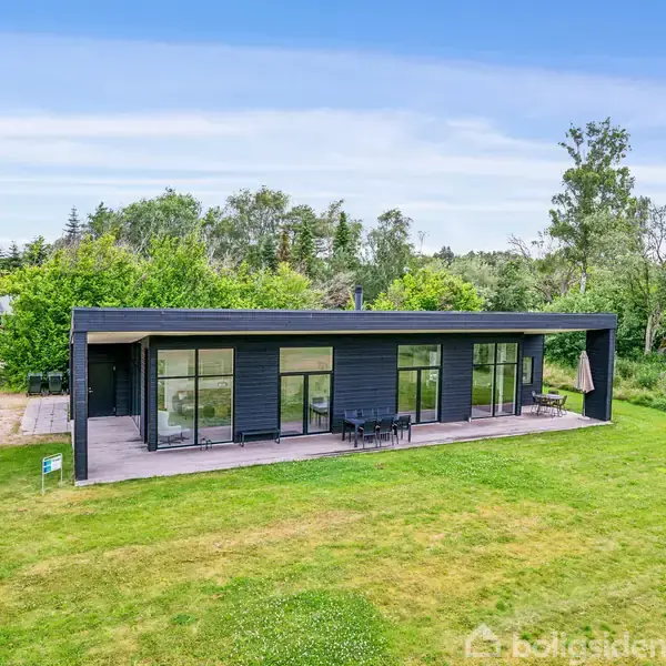 Moderne sort hus med store vinduer står stille på en græsplæne omgivet af træer og buske. Huset har en møbleret terrasse og ligger i et skovområde.