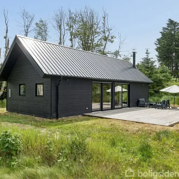 Sort træhus med moderne design står på en græsplæne. Der er en stor terrasse med havemøbler og parasol. Baggrunden er skovfyldt med høje træer.