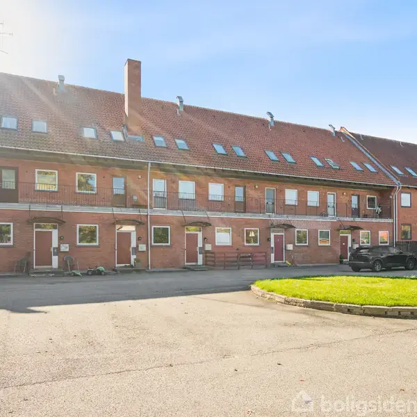 En række af røde murstensbygninger med hvide vinduer og døre, stående stille i sollys. Foran er en asfalteret plads med en rund grøn ø med græs.
