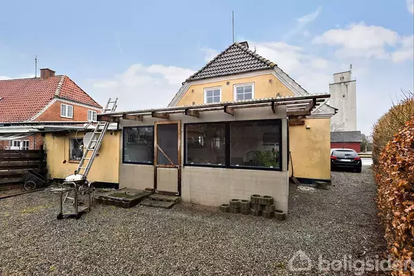 Et gult hus med skråt tag har en stige lænet op ad sig. Et værksted ses ved siden af. Huset er omgivet af grus og en bil er parkeret i baggrunden.