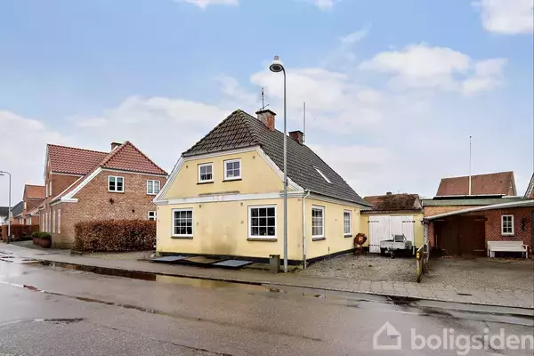 Et gult hus med sort tag står på en byvej. Der er en gadelampe foran, og andre huse ses i baggrunden under en blå himmel med skyer.