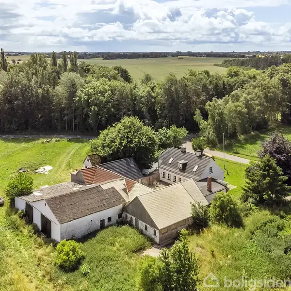 Gård med flere bygninger står stille på en grøn mark, omgivet af træer, under en overskyet himmel. En snoet vej passerer i nærheden gennem det landlige landskab.