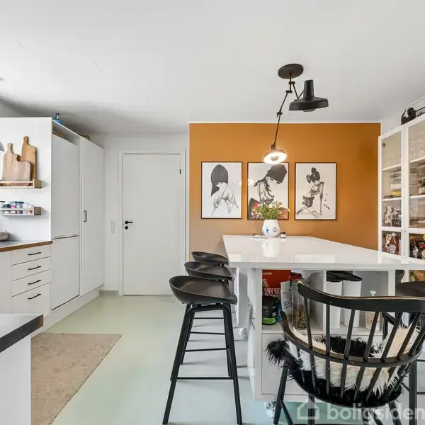Et moderne køkken har en hvid ø omgivet af sorte stole. På væggen bagved hænger to kunstplakater over en orange accentvæg. Store vinduer giver masser af naturligt lys.