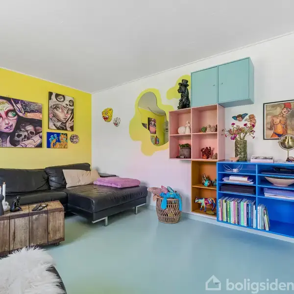 En stue med en sort lædersofa står mod en gul væg dekoreret med farverige kunstværker. En træbænk er foran sofaen. Modsat er farverige hylder med bøger, vaser og andre dekorative genstande.