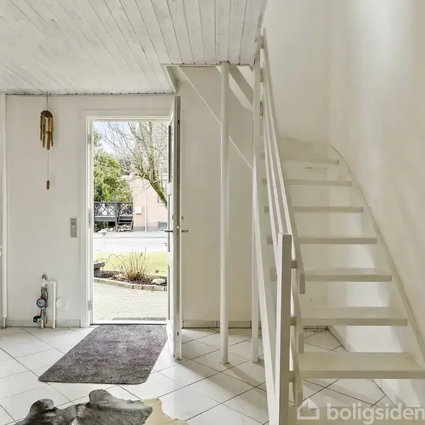 En hvid trappe fører opad i et lyst rum med flisegulv. En åben dør giver udsigt til en indkørsel. Tæpper og møbler skaber en hyggelig atmosfære.