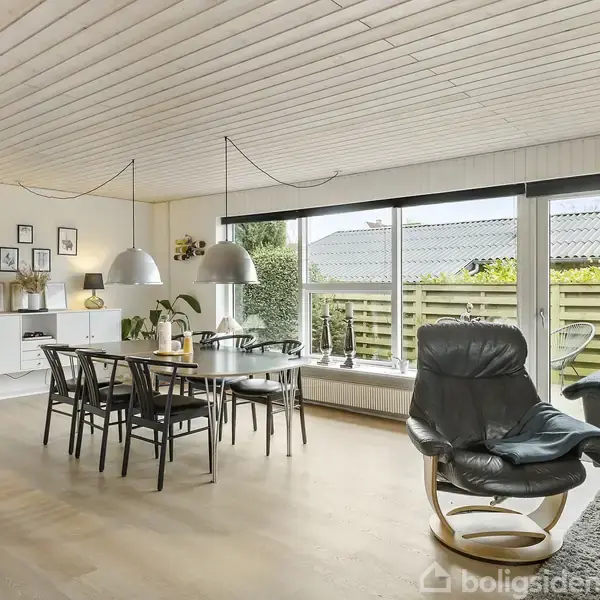 Spisebord med sorte stole står i en stue med lyse trægulve og hvidt loft. Store vinduer mod haven slipper lys ind. En sort lænestol står ved siden af sofaen.