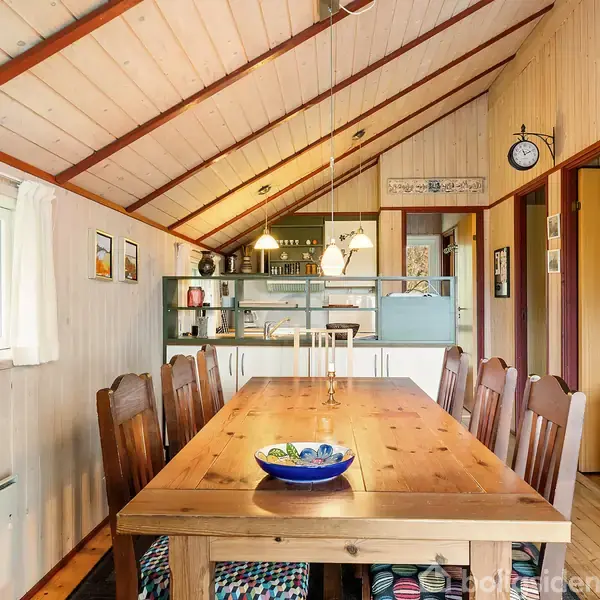 Langt spisebord med otte stole omkring. Bordet har en blå skål i midten. Omgivet af træpanelvægge og loft, vindue til venstre, køkkenområde i baggrunden.