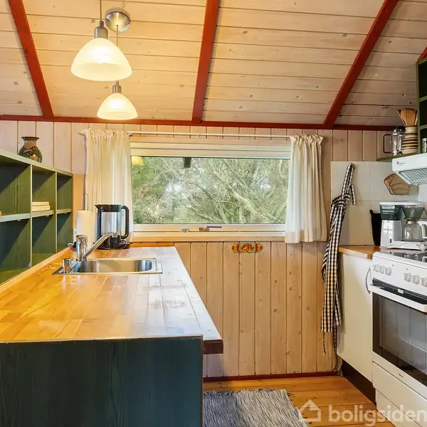 Et køkken med grønne skabe indeholder en mikrobølgeovn, kaffemaskine, og ovn. Et vindue med gardiner giver udsigt til træer. Loftet er træbeklædt med røde bjælker.