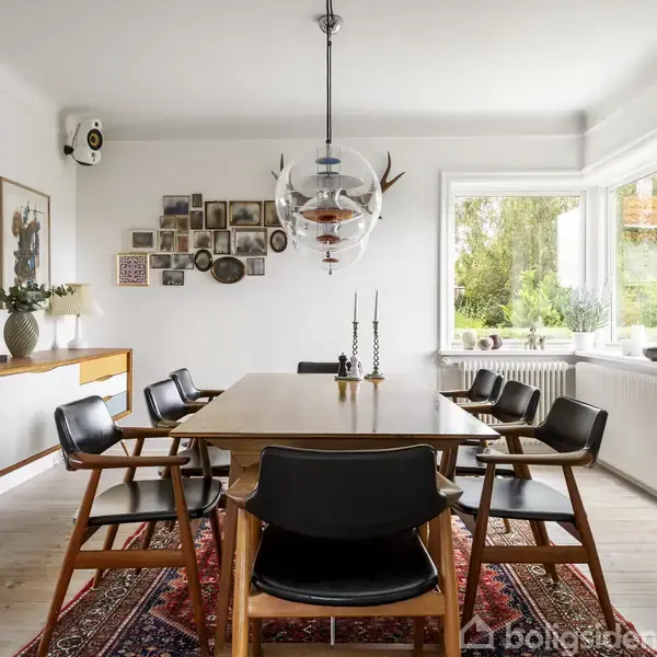 Spisebord omgivet af otte stole står på et rødt tæppe i et lyst rum. En moderne lysekrone hænger over bordet, væggen bagved prydes af kunst.