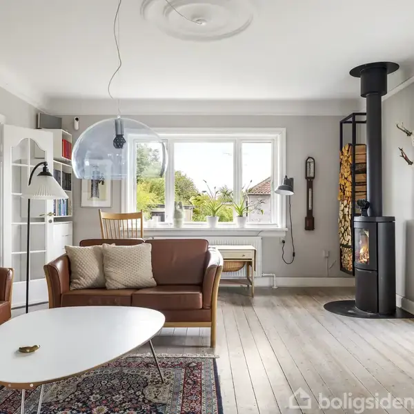Stue med brændeovn til højre, to lædersofaer omkring hvidt sofabord. Store vinduer lukker lys ind. Hylder og lampe til venstre. Trægulv og tæppe under bordet.