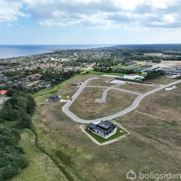 Et moderne hus står i et åbent landskab nær en snoet vej; omgivet af marker og med en udsigt over en by og havet i horisonten under en skyet himmel.