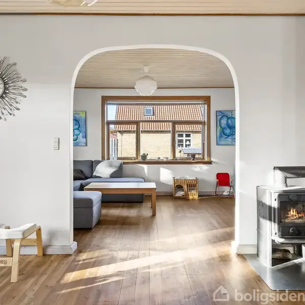 En lyseblå sofa står stille i en stue, omgivet af lyse træmøbler og dekorationer. Store vinduer slipper lys ind, mens malerier og en lampe pynter væggene.