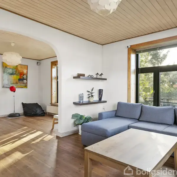 En blå sofa står i en lys stue, omgivet af et træbord, reoler med dekorationer, store vinduer og kunst på væggene. Sollys strømmer ind, hvilket giver en hyggelig atmosfære.